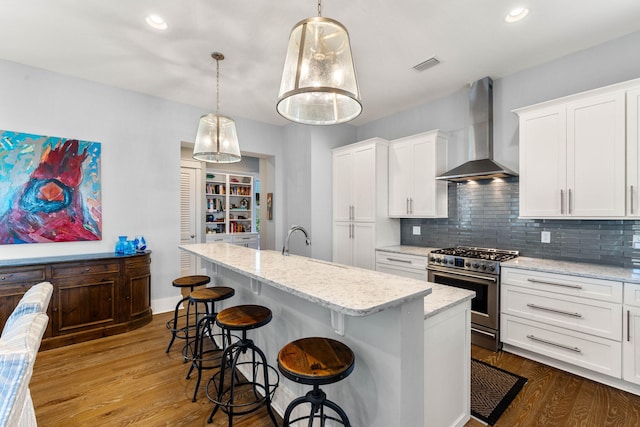 kitchen featuring wall chimney range hood, high end stove, an island with sink, pendant lighting, and wood-type flooring