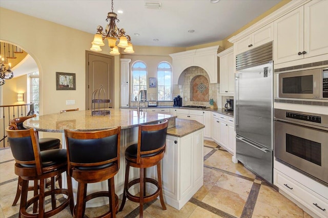 kitchen featuring built in appliances, light stone countertops, a center island with sink, and an inviting chandelier