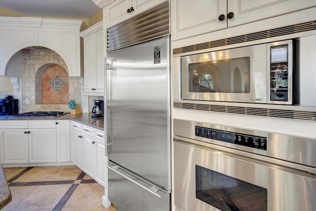 kitchen with white cabinets, built in appliances, decorative backsplash, light tile patterned floors, and custom range hood