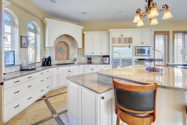 kitchen featuring a kitchen bar, tasteful backsplash, built in appliances, hanging light fixtures, and an island with sink