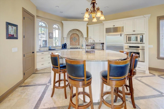 kitchen featuring decorative backsplash, light stone countertops, a kitchen island with sink, built in appliances, and hanging light fixtures