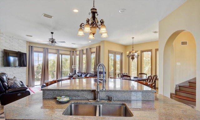 kitchen with sink, hanging light fixtures, light stone counters, a kitchen island with sink, and ceiling fan with notable chandelier