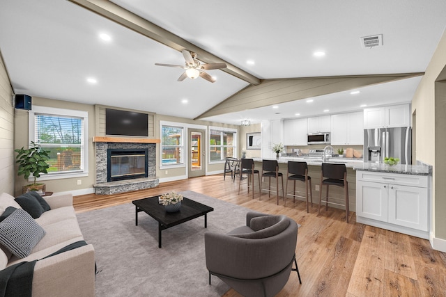 living room with vaulted ceiling with beams, ceiling fan, light hardwood / wood-style flooring, and a stone fireplace