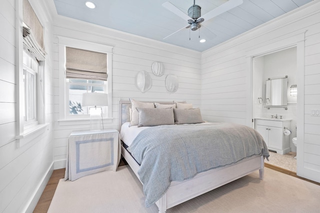 bedroom with ensuite bath, ceiling fan, light hardwood / wood-style flooring, and wood walls