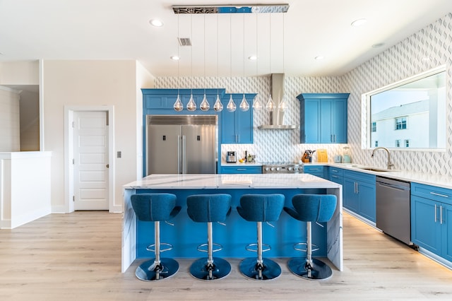 kitchen featuring light wood-type flooring, stainless steel appliances, wall chimney range hood, a center island, and hanging light fixtures