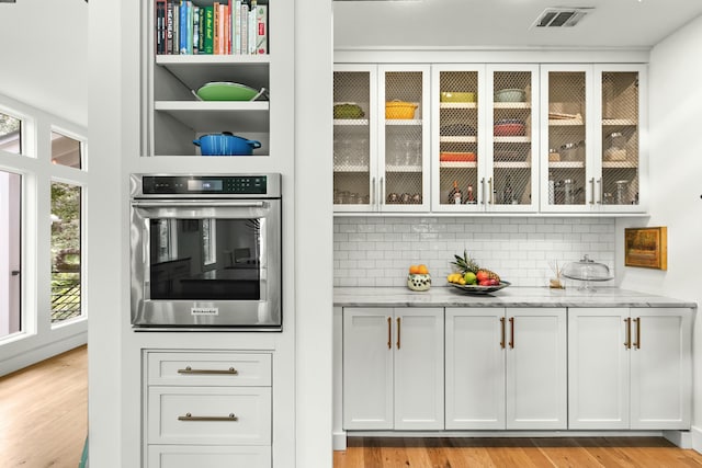 interior space featuring backsplash, light stone counters, stainless steel oven, light hardwood / wood-style flooring, and white cabinetry