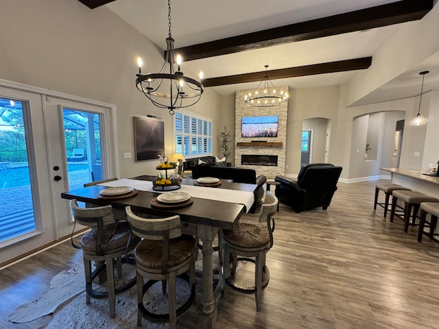 dining room featuring beamed ceiling, wood-type flooring, a chandelier, and a stone fireplace