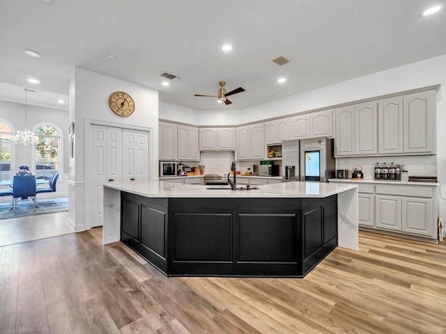 kitchen featuring stainless steel refrigerator with ice dispenser, ceiling fan with notable chandelier, sink, light hardwood / wood-style floors, and an island with sink