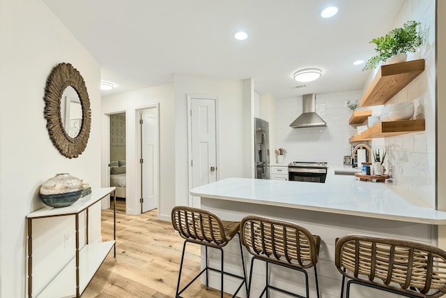 kitchen with a kitchen breakfast bar, wall chimney range hood, light hardwood / wood-style flooring, appliances with stainless steel finishes, and kitchen peninsula