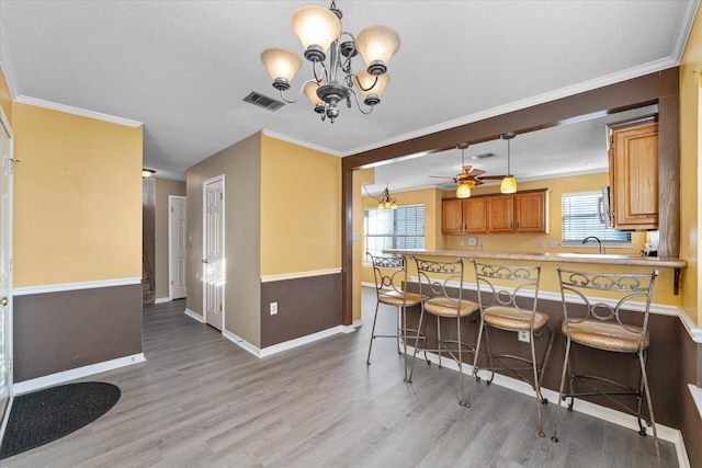 kitchen with ceiling fan with notable chandelier, light wood-type flooring, ornamental molding, kitchen peninsula, and a breakfast bar area