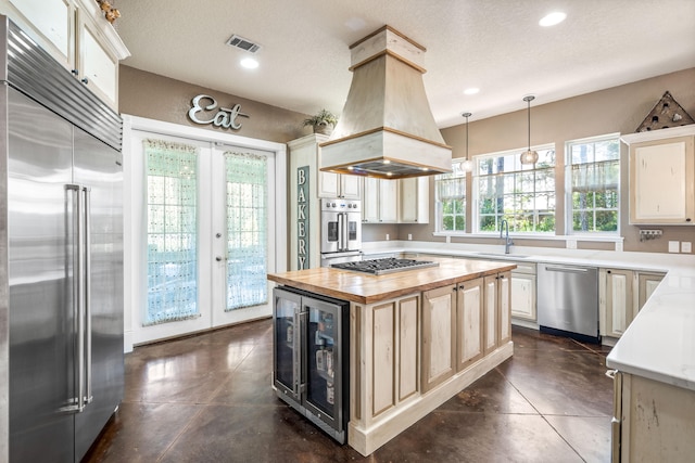 kitchen featuring pendant lighting, appliances with stainless steel finishes, a kitchen island, custom range hood, and beverage cooler