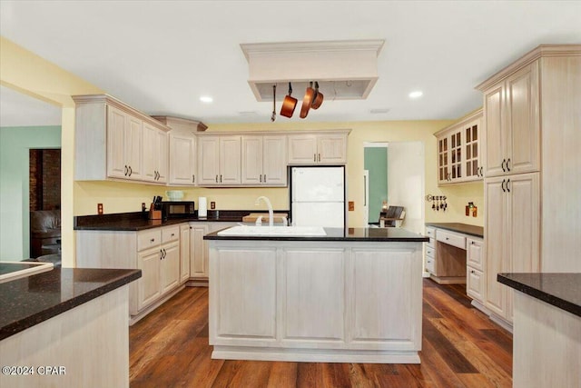 kitchen with cream cabinetry, white fridge, dark wood-type flooring, and an island with sink