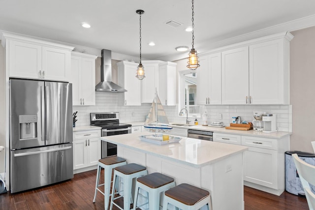 kitchen featuring dark wood-type flooring, wall chimney exhaust hood, appliances with stainless steel finishes, decorative light fixtures, and white cabinetry