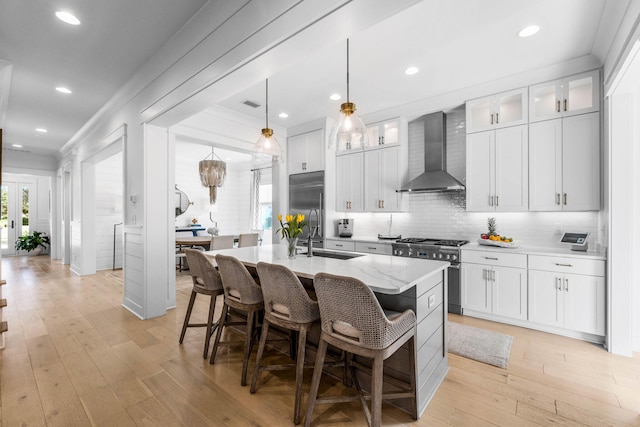 kitchen with high end appliances, sink, wall chimney exhaust hood, light wood-type flooring, and an island with sink