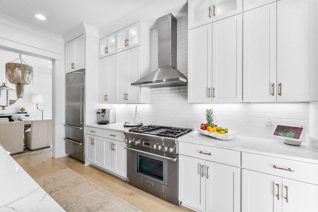 kitchen with high end appliances, white cabinets, wall chimney exhaust hood, light hardwood / wood-style floors, and light stone counters