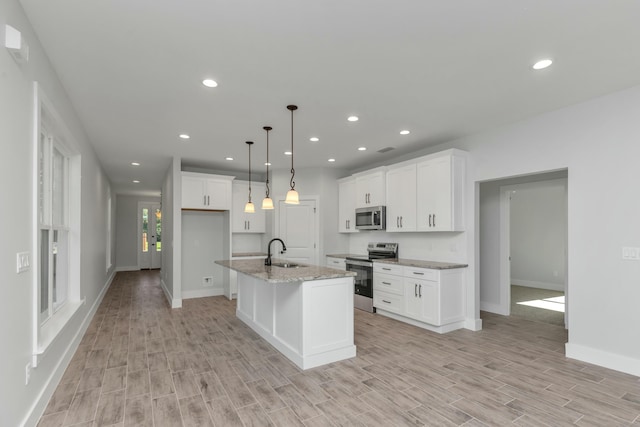 kitchen with light stone countertops, stainless steel appliances, decorative light fixtures, white cabinets, and an island with sink