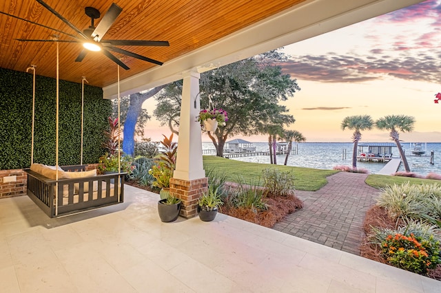 patio terrace at dusk featuring ceiling fan, a yard, and a water view