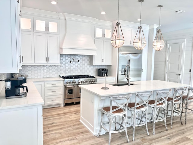 kitchen featuring pendant lighting, premium range hood, an island with sink, premium appliances, and white cabinetry