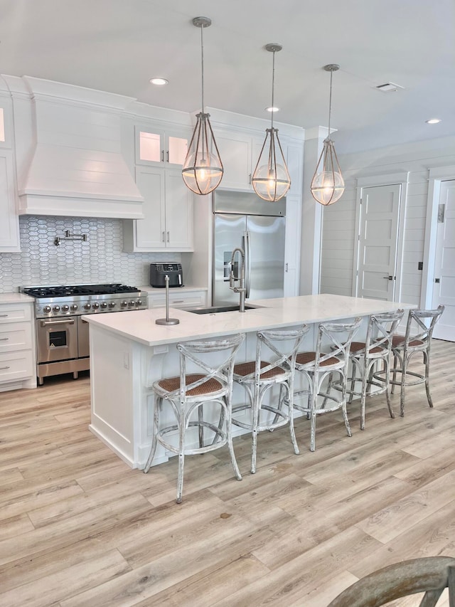 kitchen with custom exhaust hood, high end appliances, pendant lighting, a center island with sink, and white cabinets