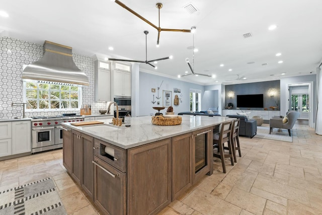 kitchen with premium range hood, light stone counters, double oven range, white cabinets, and an island with sink