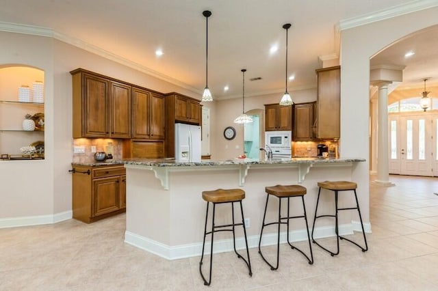 kitchen featuring white appliances, light stone countertops, ornate columns, a peninsula, and brown cabinets