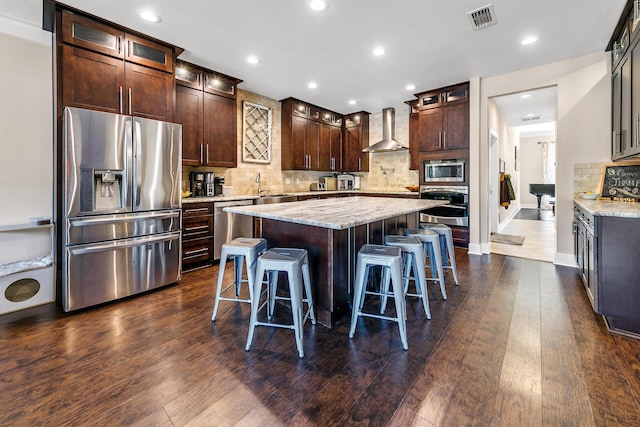 kitchen with light stone countertops, dark hardwood / wood-style flooring, wall chimney exhaust hood, stainless steel appliances, and a kitchen island