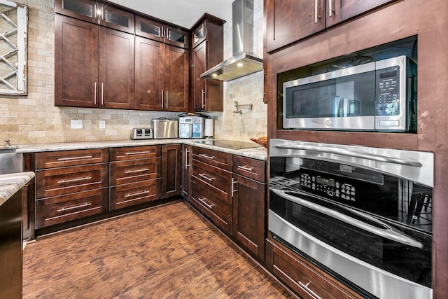 kitchen featuring wall chimney exhaust hood, light stone counters, dark hardwood / wood-style flooring, backsplash, and appliances with stainless steel finishes