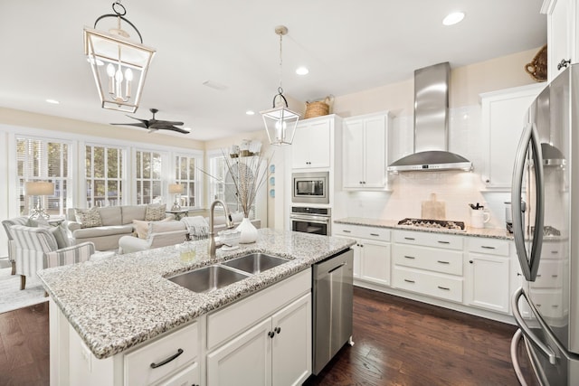 kitchen with appliances with stainless steel finishes, wall chimney exhaust hood, sink, ceiling fan, and a center island with sink