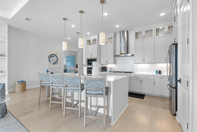 kitchen with an island with sink, stainless steel appliances, white cabinetry, and wall chimney range hood