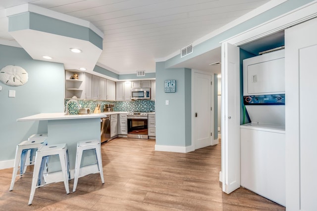 kitchen with light hardwood / wood-style flooring, kitchen peninsula, stacked washer / drying machine, a kitchen bar, and appliances with stainless steel finishes