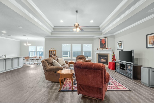 living room with ceiling fan with notable chandelier, a tray ceiling, ornamental molding, and a tiled fireplace