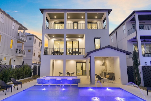 back house at dusk featuring a swimming pool with hot tub, a patio area, a balcony, and pool water feature