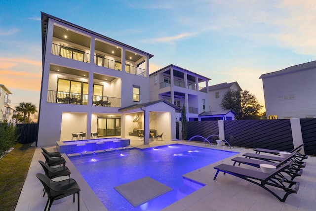 back house at dusk with a patio area, a pool with hot tub, a balcony, and pool water feature