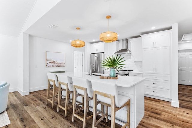 kitchen with wall chimney exhaust hood, stainless steel appliances, an island with sink, pendant lighting, and white cabinets