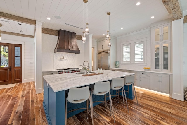 kitchen with dark wood-type flooring, plenty of natural light, a center island with sink, custom range hood, and appliances with stainless steel finishes