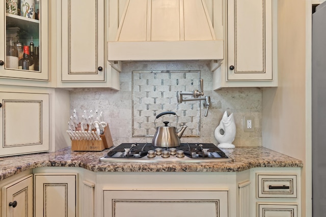 kitchen featuring stainless steel gas stovetop, custom exhaust hood, backsplash, cream cabinets, and stone countertops