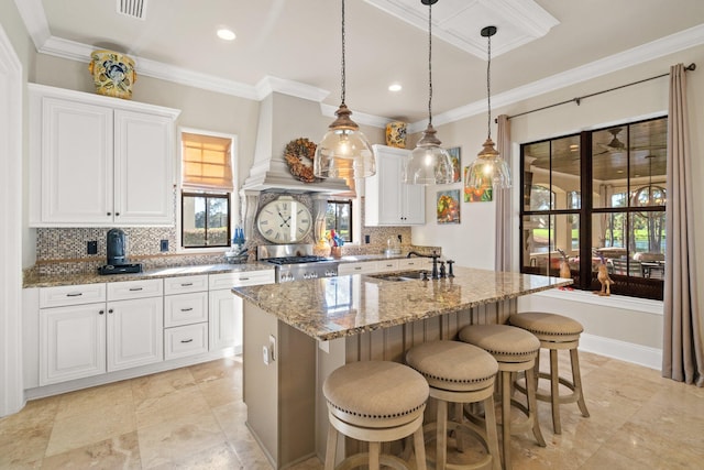 kitchen featuring white cabinetry, light stone countertops, decorative light fixtures, a center island with sink, and custom exhaust hood