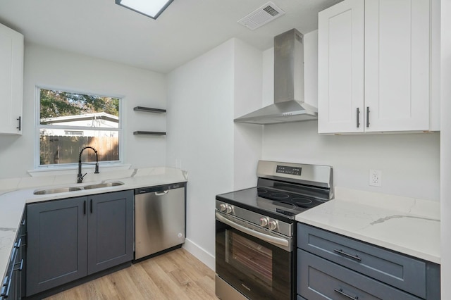 kitchen with wall chimney range hood, sink, light wood-type flooring, appliances with stainless steel finishes, and white cabinetry