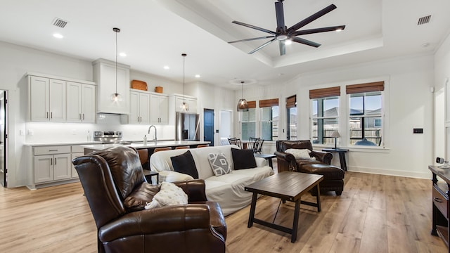 living room featuring crown molding, sink, ceiling fan, light wood-type flooring, and a tray ceiling