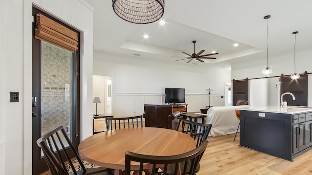 dining space with ceiling fan, sink, a barn door, a tray ceiling, and light wood-type flooring