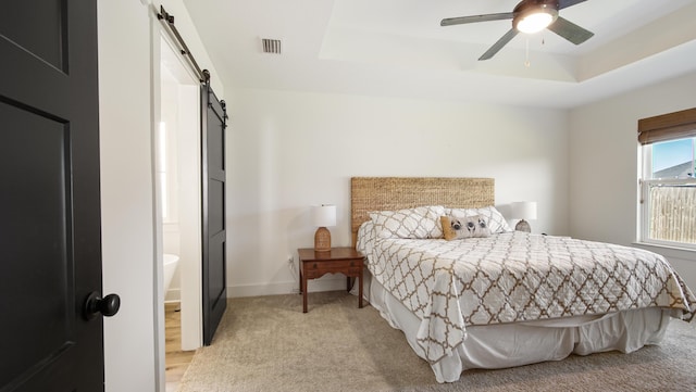 carpeted bedroom with a barn door, ensuite bathroom, a raised ceiling, and ceiling fan