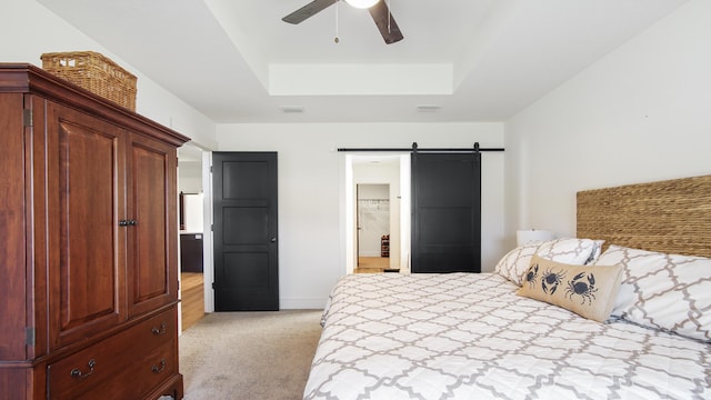 bedroom featuring ceiling fan, a raised ceiling, a barn door, ensuite bathroom, and light carpet