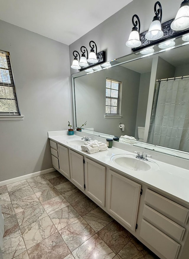 bathroom featuring toilet, a sink, baseboards, marble finish floor, and double vanity