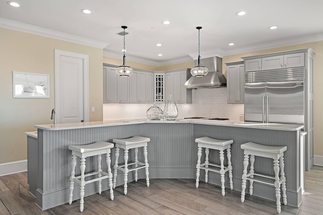 kitchen featuring a kitchen bar, built in refrigerator, gray cabinetry, pendant lighting, and wall chimney exhaust hood