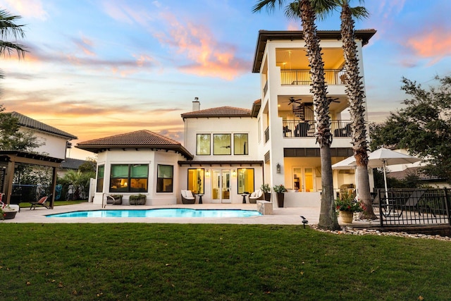 back house at dusk with a fenced in pool, a patio area, a balcony, and a lawn