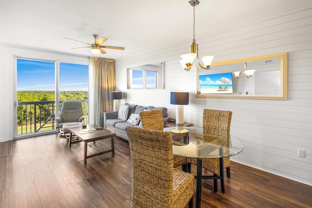 dining space with ceiling fan with notable chandelier, dark wood-type flooring, and wood walls