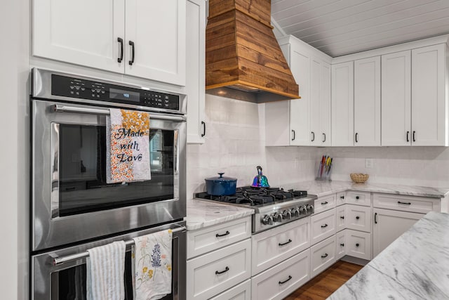 kitchen featuring white cabinets, appliances with stainless steel finishes, custom exhaust hood, decorative backsplash, and light stone counters