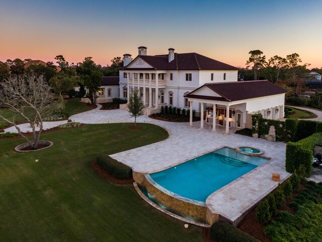 back house at dusk featuring a lawn, a patio area, a swimming pool with hot tub, and a balcony