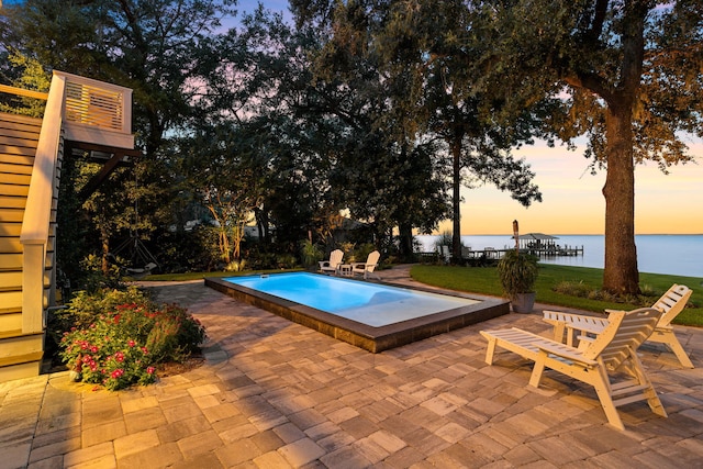 pool at dusk featuring a patio area, a water view, and a yard