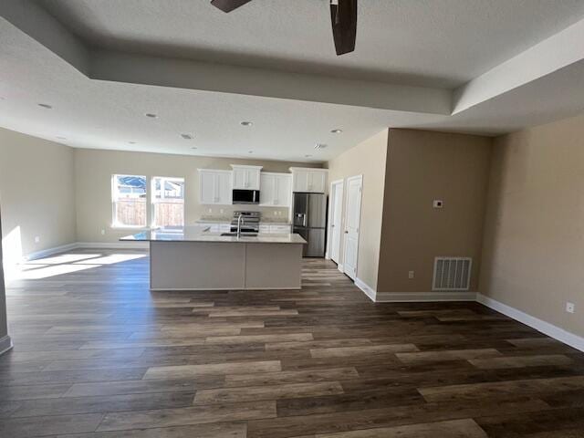 kitchen with dark wood-type flooring, white cabinetry, appliances with stainless steel finishes, a raised ceiling, and a kitchen island with sink